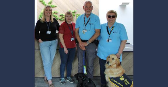 4 People Standing In A Row With A Blonde Guide Dog And A Black Guide Dog