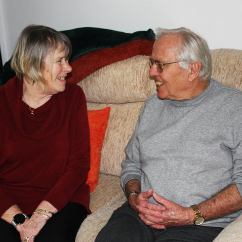 An Older Couple Who Live At Swallowtail Place, Sitting On A Sofa Smiling At Each Other